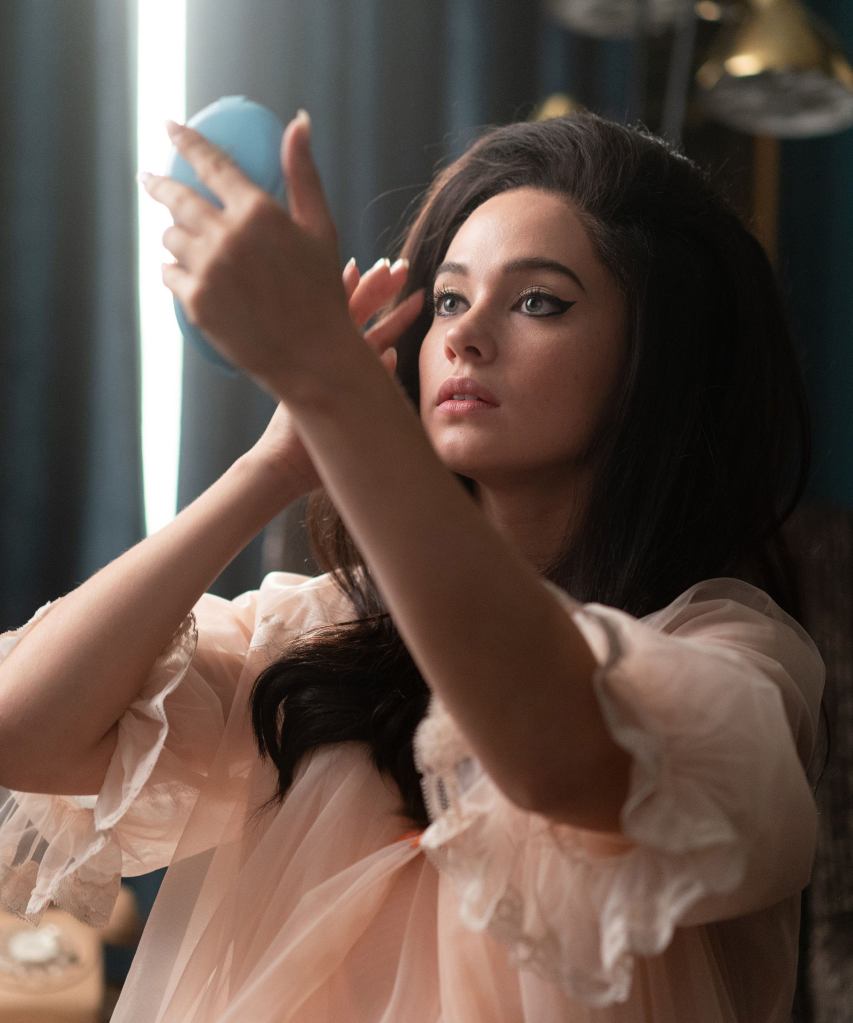 Priscilla holds a blue hand mirror while studying her makeup in a pink nightgown.