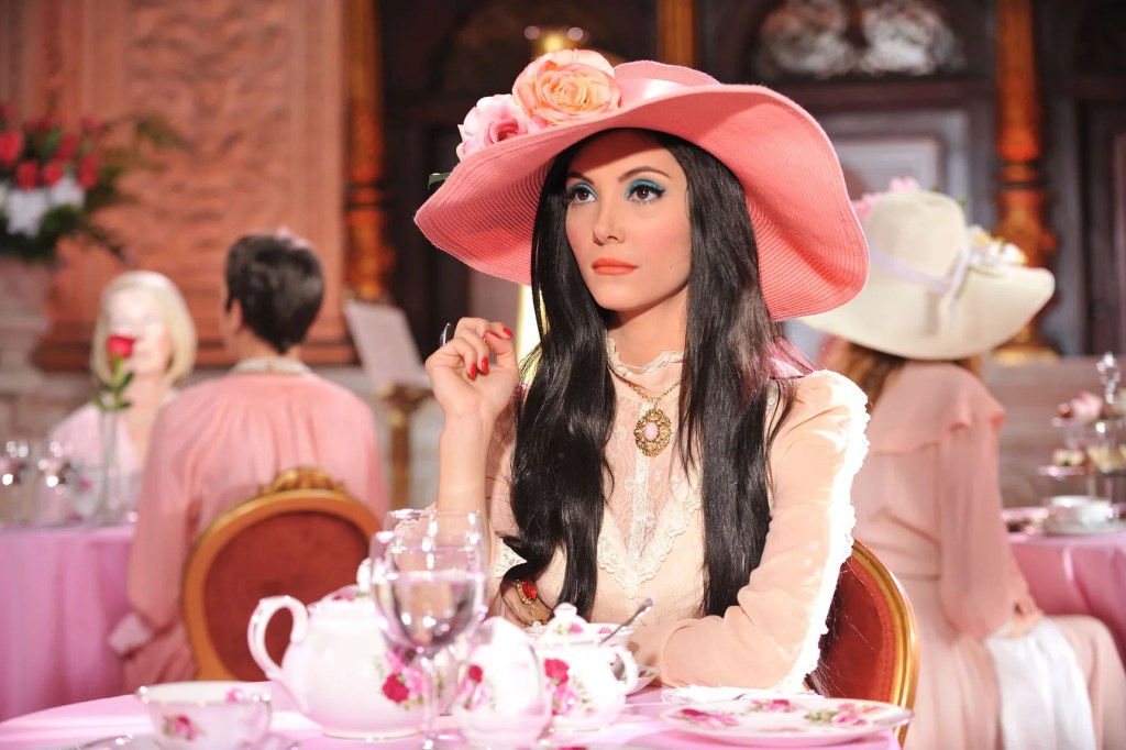 Elaine sits in a pink tearoom wearing a flowered hat, lace clothing, blue eyeshadow, and red nails.