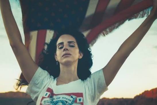 Lana Del Rey stands beneath an American flag with her arms raised and eyes closed against a desert landscape.