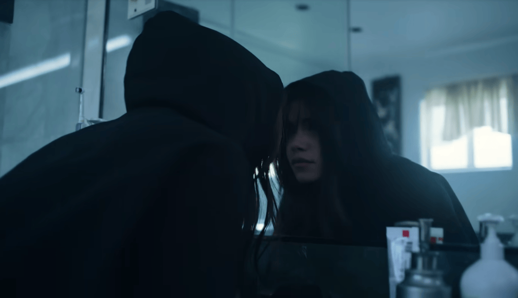 Nessa Barrett faces a bathroom mirror wearing a black hood while cold blue light fills the room.